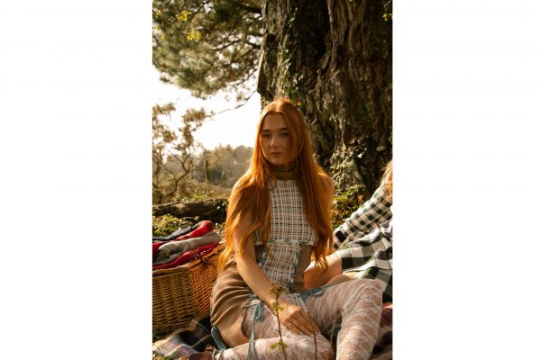 A photograph of a lone figure sat on a picnic blanket wearing a tartan knitted dress with a knitted roll neck and lace stockings.
